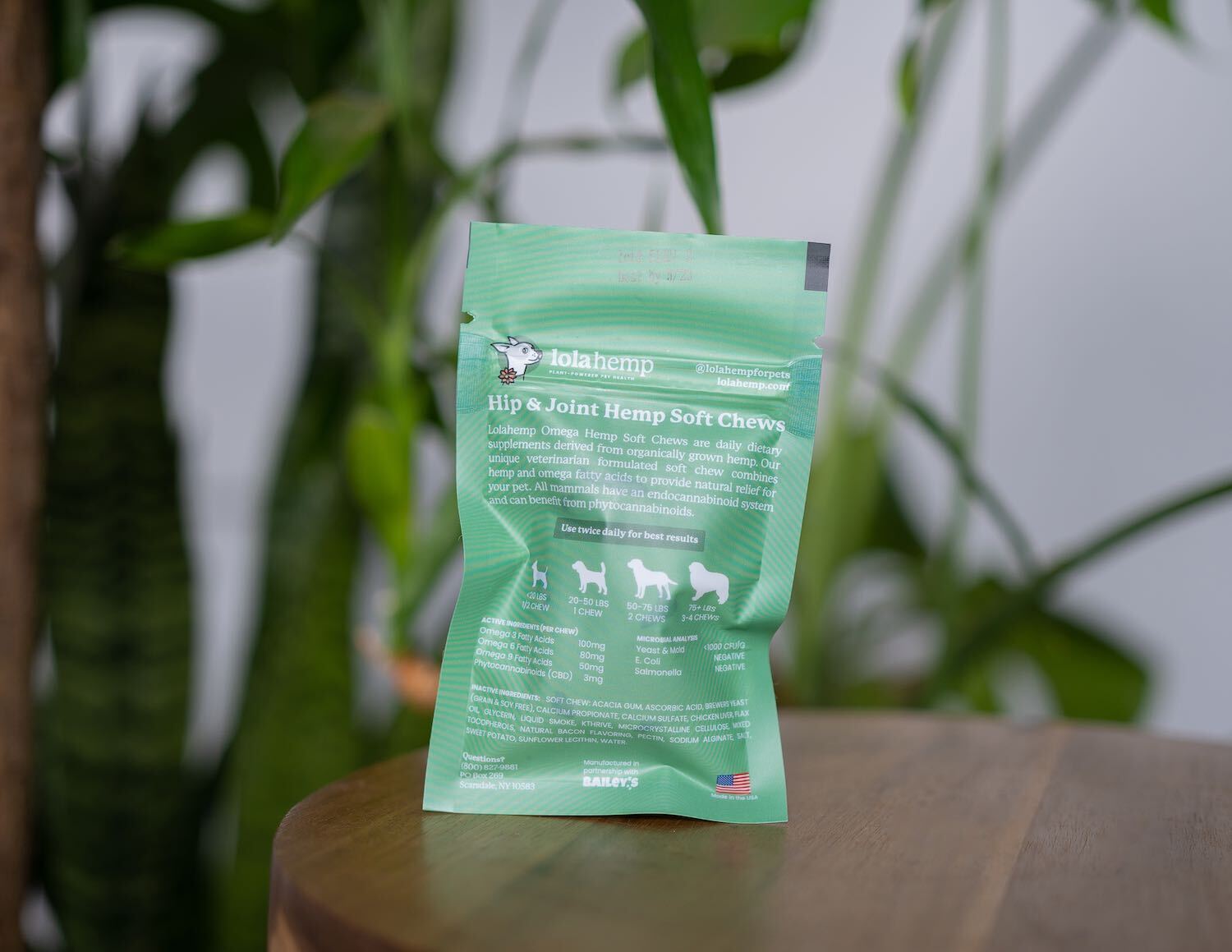 back of mini green colored bag of regular strength lolahemp hip & joint hemp soft chews on wooden table with plants in the background