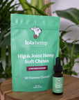 Front of green colored bag of regular strength lolahemp hip & joint hemp soft chews with brown 150mg lolahemp oil bottle with green label on wooden table with plants in the background