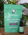 Front of green colored bag of regular strength lolahemp hip & joint hemp soft chews with brown 300mg lolahemp oil bottle with green label on wooden table with plants in the background