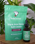 Front of green colored bag of regular strength lolahemp hip & joint hemp soft chews with brown 600mg lolahemp oil bottle with green label on wooden table with plants in the background