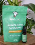 Green colored bag of regular strength lolahemp calming hemp soft chews with brown 150mg regular strength lolahemp oil bottle with green label on wooden table with plants in the background