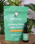 Green colored bag of regular strength lolahemp calming hemp soft chews with brown 300mg regular strength lolahemp oil bottle with green label on wooden table with plants in the background