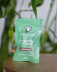 Front of mini green colored bag of regular strength lolahemp calming hemp soft chews on wooden table with plants in the background